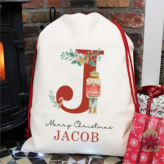 Linen Hessian Christmas Santa sack with red strings picturing a red Initial embellished with either a nutcracker or a plum fairy. Underneath the initial there is merry christmas and the boy or girl&#39;s name.